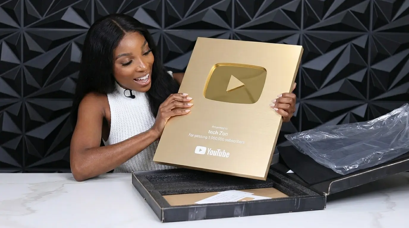 PROMPT: A close-up lifestyle photograph of a creator's hands carefully unboxing a YouTube Gold Play Button from its custom presentation box. The gold-plated brass plaque catches warm studio lighting, showing the mirror-like gold finish and laser-etched channel name. The background is a blurred modern studio setup with ring lights and monitors. Photorealistic, warm color tones.