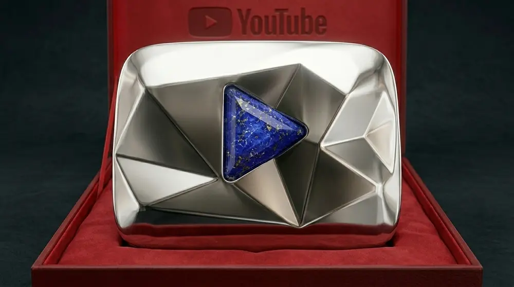 PROMPT: A dramatic side-by-side comparison photograph showing the progression of four YouTube play button awards on a dark museum-style shelf. From left to right: a yellow-orange Tiger's Eye award, a green Malachite award, a blue Lapis Lazuli award, and a massive custom silver Beast Panther trophy. Each award is dramatically lit from above with its own coloured accent light matching its gemstone. Ultra-high detail commercial photography.