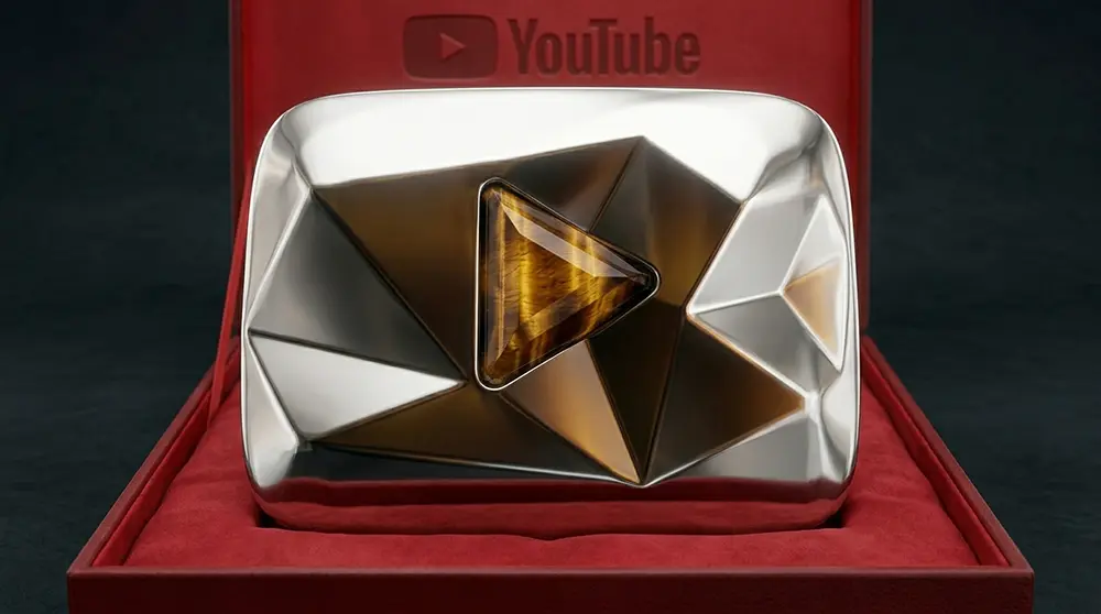 PROMPT: A dramatic product photograph showing three YouTube Tiger's Eye play button awards displayed in a row on a dark reflective surface. Each award features a heavy polished steel casing with a vivid yellow-orange crystal centrepiece shaped like the YouTube play button triangle. Warm amber-toned studio lighting from above creates golden reflections. Ultra-high detail commercial photography with a dark moody background.